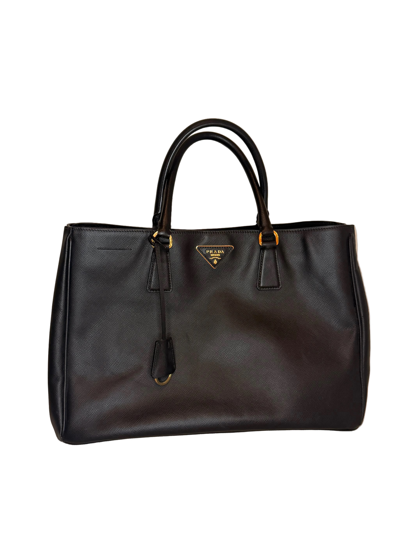 A black leather Prada Saffiano tote bag with gold-tone hardware, displayed on a light beige textured surface against a solid beige background.