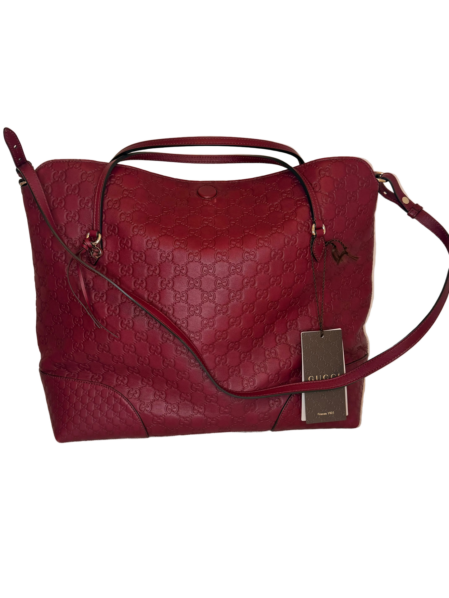A burgundy red Gucci leather handbag with the brand's signature GG monogram pattern, featuring dual top handles, a detachable shoulder strap, and a hanging brand tag, displayed on a light-colored fabric-covered surface.