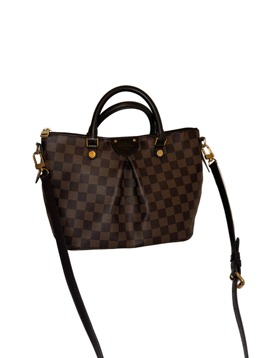A Louis Vuitton handbag in the Damier Ebene checkered pattern, featuring dark brown and black checkered squares, black leather handles and shoulder strap, and gold-tone hardware, displayed on a light beige textured surface against a neutral beige background.