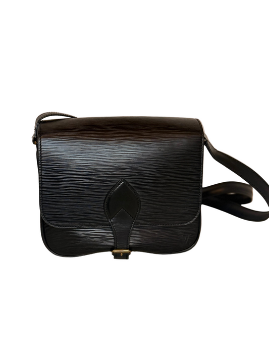 A black textured leather shoulder bag with a central strap detail and a small gold-tone buckle, resting on a light beige fabric surface against a plain beige background.