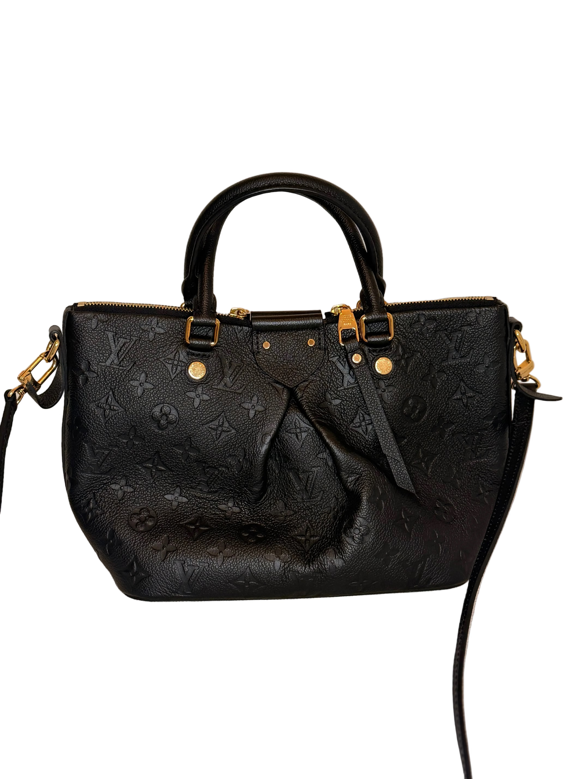 A black Louis Vuitton monogram leather handbag with gold-tone hardware, featuring dual top handles and a shoulder strap, displayed on a light beige fabric surface against a plain beige wall.
