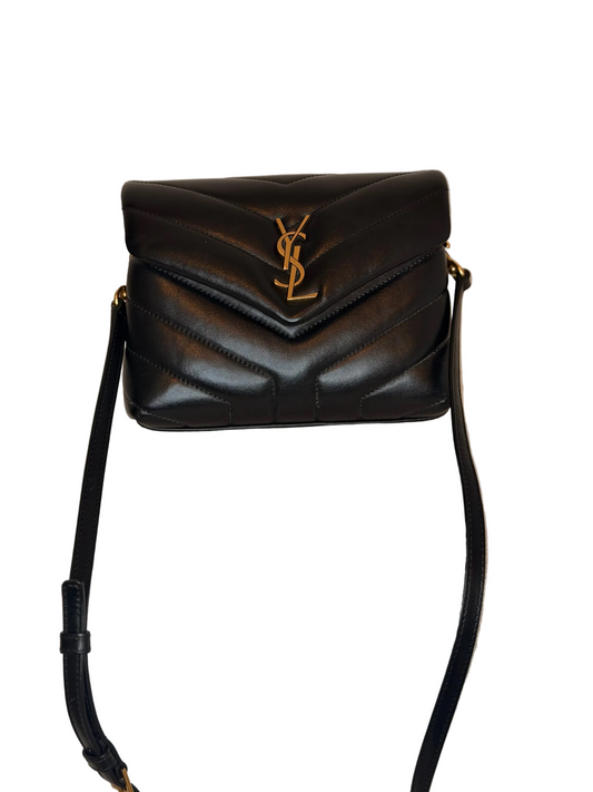 A black leather shoulder bag with a gold-tone YSL monogram clasp, resting on a light beige textured fabric surface against a neutral beige background.