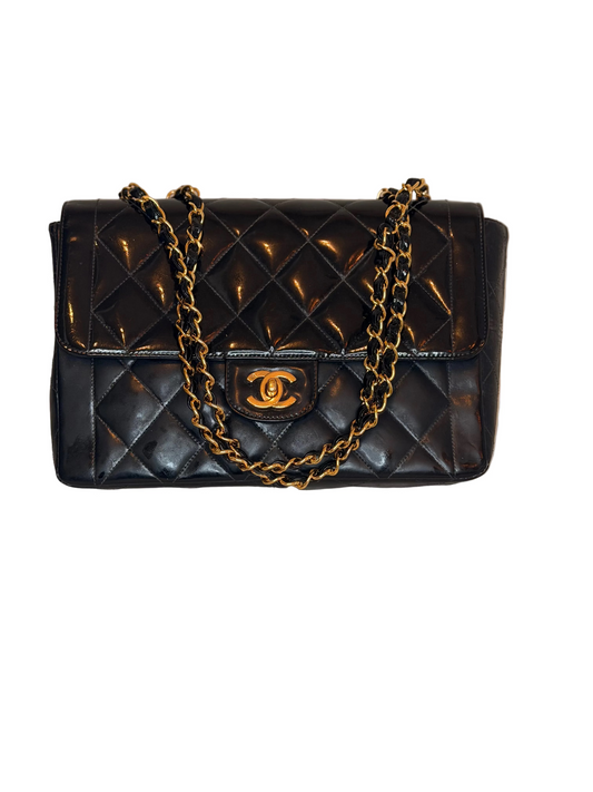 A black quilted leather handbag with a gold-tone chain strap and a gold interlocking CC logo on the front, displayed on a light beige textured surface against a neutral beige background.