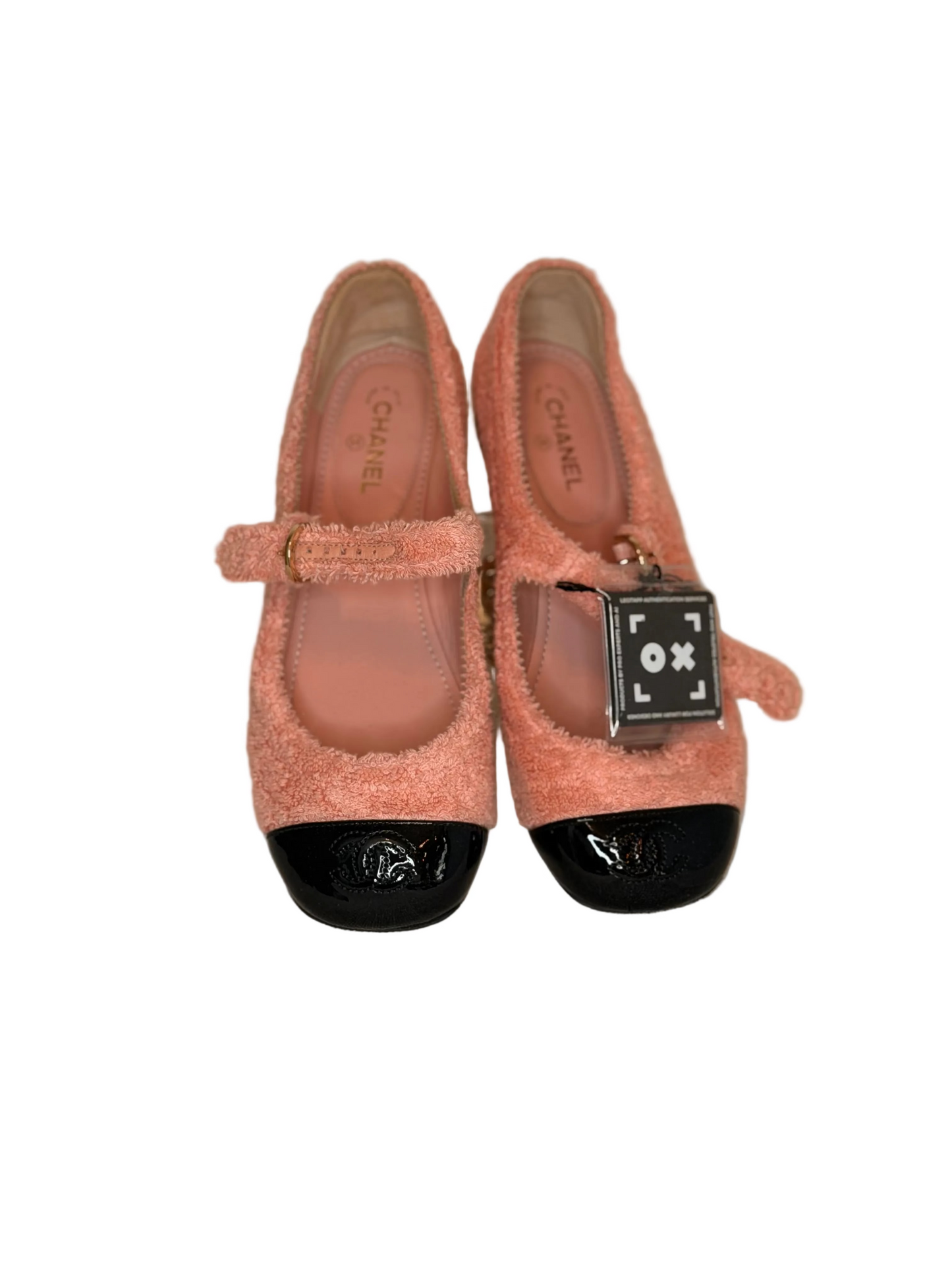 A pair of Chanel Mary Jane shoes in a textured peach-pink fuzzy fabric with black patent leather toe caps and a signature black CC logo, displayed on a neutral beige fabric surface.